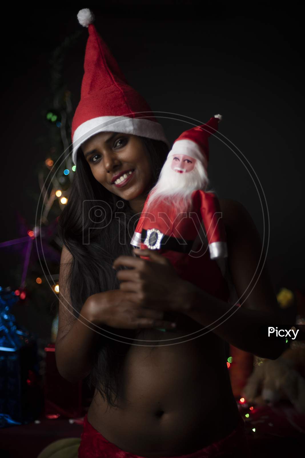 Portrait of young Indian Bengali dark skinned brunette woman in Santa Clause lingerie, gifts and soft toys in decorative Christmas tree background.Indian lifestyle and Christmas celebration.