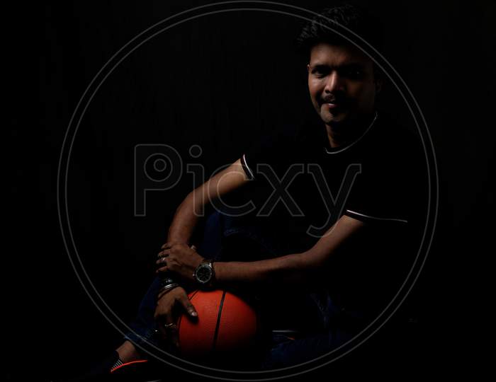 Portrait of a handsome and intelligent Indian brunette man wearing a solid black t shirt standing before a copy space black background holding a basketball. Indian lifestyle and fashion portrait