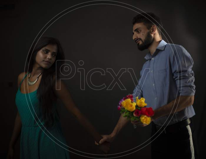 A Kashmiri/European/Arabian man in formal wear trying to give flowers to his dark skinned Indian/African girlfriend/wife in front of a black copy space studio background. lifestyle and fashion