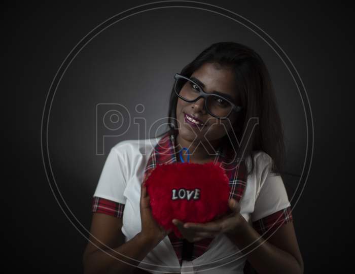 Portrait of an Asian/Indian/African brunette dark skinned young girl in sexy school uniform and spectacles holding red cushion in a black/grey studio background. Fashion and cosplay photography.