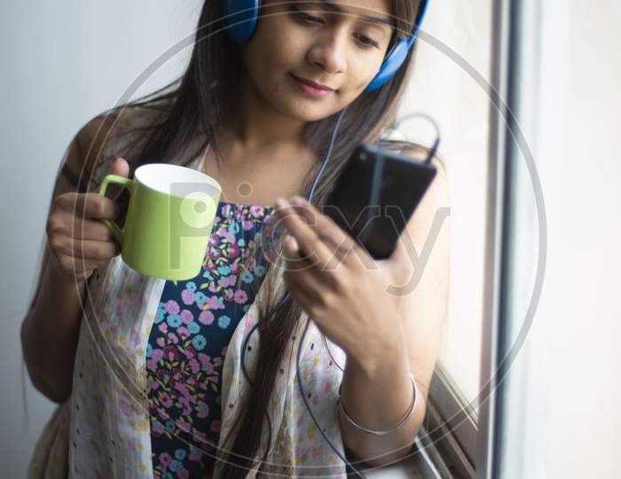 An young and attractive Indian  brunette woman in white sleeping wear listening to music through headphone on her mobile standing with a coffee near the window in white background. Indian lifestyle.