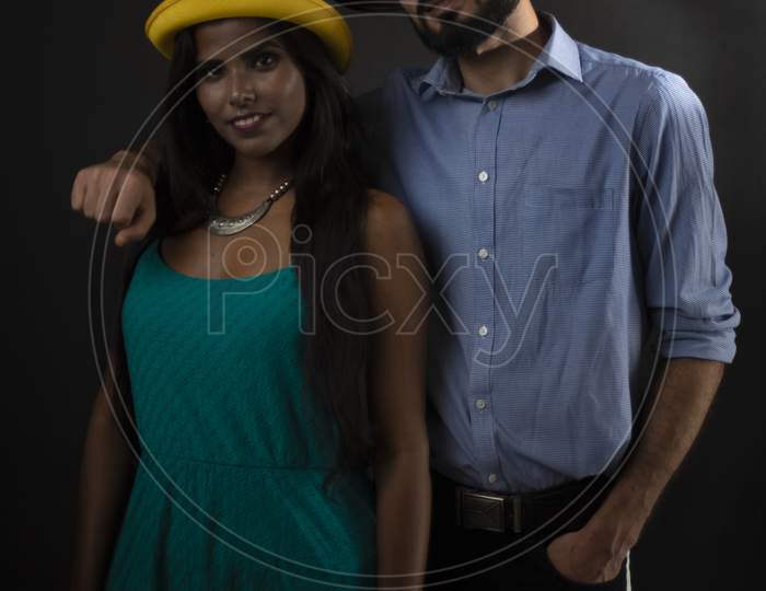A dark skinned Indian/African girl and a Kasmiri/European/Arabian man in formal wear with fashionable hat in front of a black copy space studio background. Indian lifestyle and fashion photography.