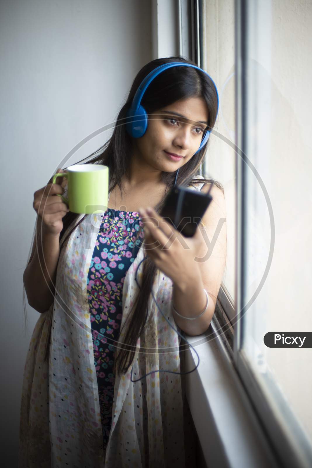 An young and attractive Indian  brunette woman in white sleeping wear listening to music through headphone on her mobile standing with a coffee near the window in white background. Indian lifestyle.