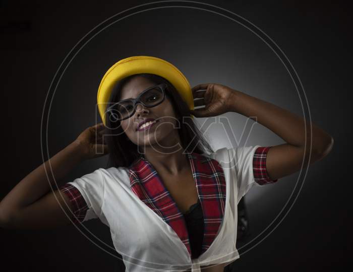 Portrait of an Asian/Indian/African brunette dark skinned young girl in sexy school uniform and spectacles with yellow hat in a black/grey studio background. Fashion and cosplay photography.