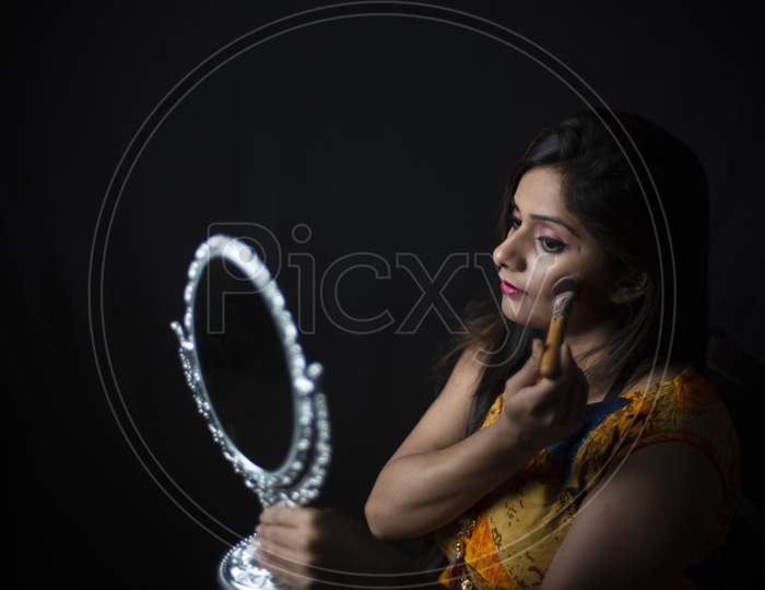 Young and attractive Indian Bengali brunette woman is looking her face in a mirror while doing her hairdo sitting in black studio copy space background. Indian lifestyle.