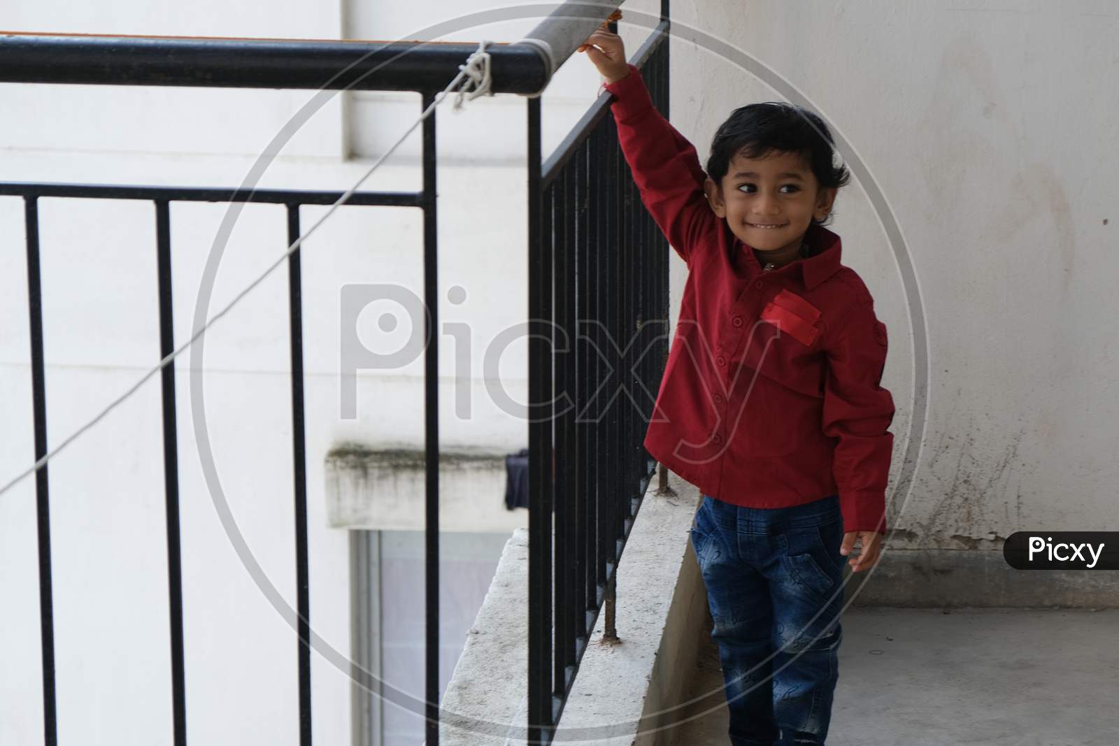 Portrait of Indian cute little cheerful brunette Tamil baby boy wearing vibrant red shirt while standing on a balcony in a white urban background. Indian lifestyle.