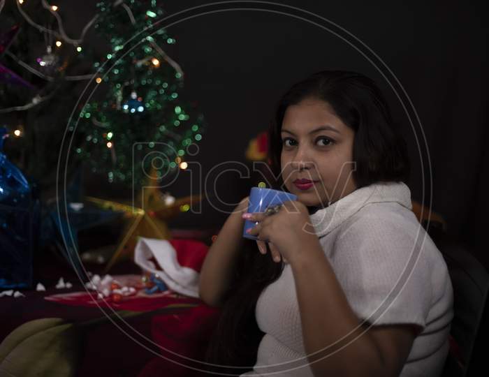 An young and attractive brunette plus size Indian Bengali woman in western woolen sweater enjoying with coffee/tea in decorative Christmas tree background.Indian lifestyle and Christmas celebration
