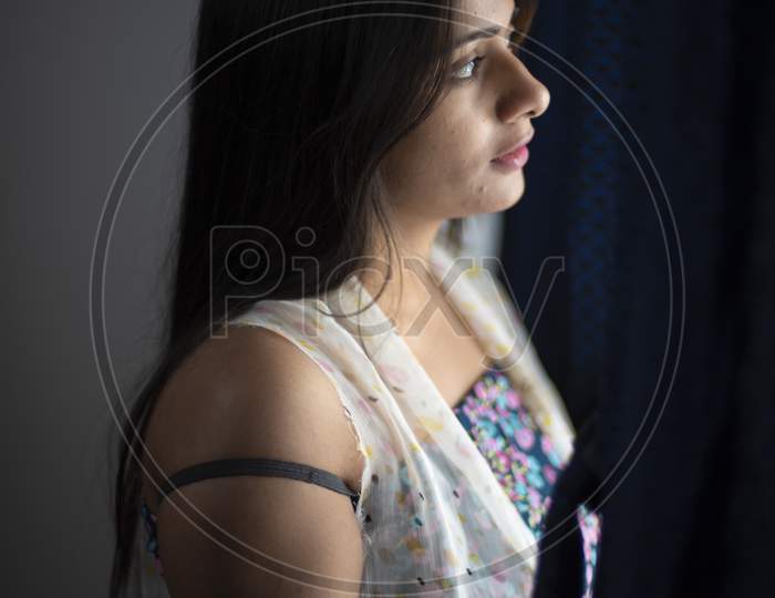 An young and attractive Indian brunette woman in white sleeping wear looking outside through the window removing the curtain in dark background. Indian lifestyle.