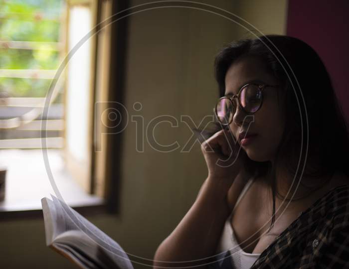 A young and attractive Indian Bengali brunette woman in white sleeping wear with a coffee/tea cup reading book on a bed in front of a window inside in her room. Indian lifestyle.