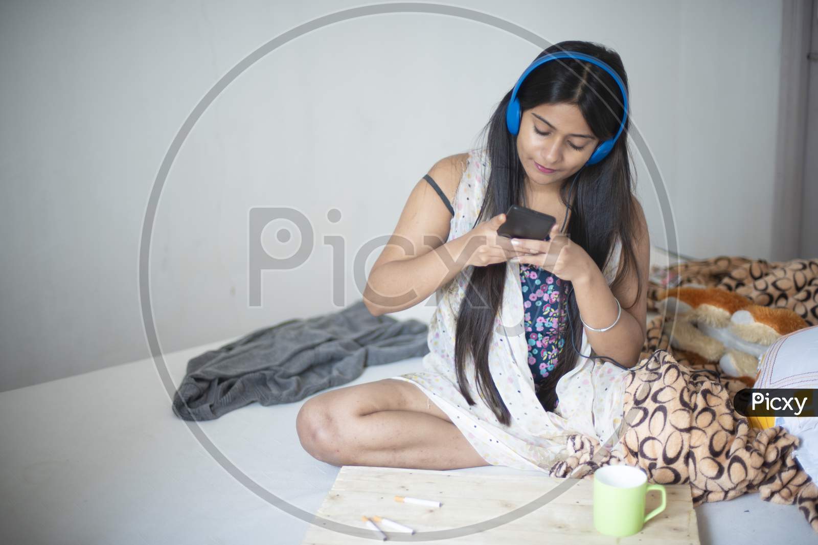 An young Indian Bengali brunette girl in white sleeping wear is listening music while sitting on bed with cellphone,coffee/tea cup and earplug in a casual mood in the winter morning. Indian lifestyle.