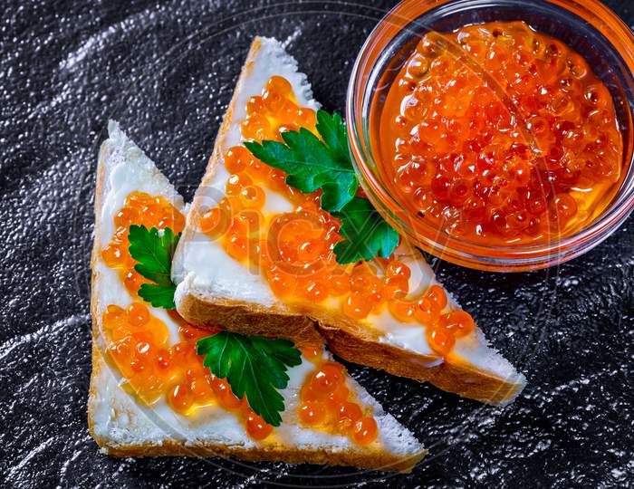 Red Caviar In A Glass Bowl And On Sandwiches On A Black Stone Background