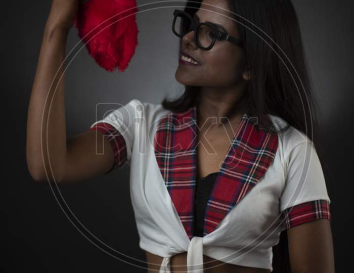 Portrait of an Asian/Indian/African brunette dark skinned young girl in sexy school uniform and spectacles holding red cushion in a black/grey studio background. Fashion and cosplay photography.