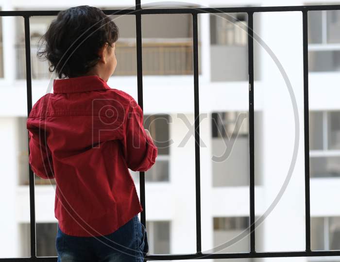 Backside portrait of Indian cute little cheerful brunette Tamil baby boy wearing vibrant red shirt while standing on a balcony in a white urban background. Indian lifestyle.