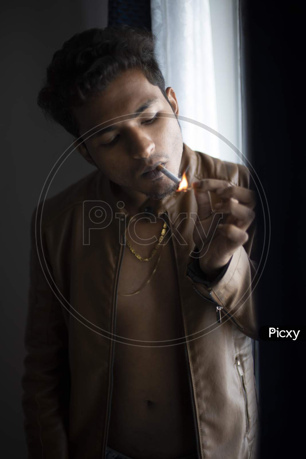 Close up portrait of an young and handsome man with front open leather jacket lightening cigarette while standing in front of a glass door of a balcony. Indian lifestyle.