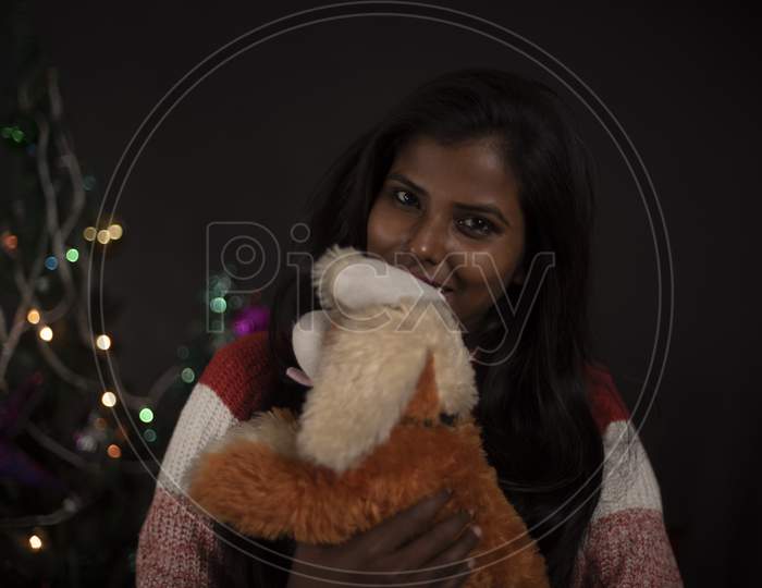 Portrait of young and attractive brunette dark skinned Indian Bengali woman in western woolen sweater with soft toy in decorative Christmas tree background.Indian lifestyle and Christmas celebration