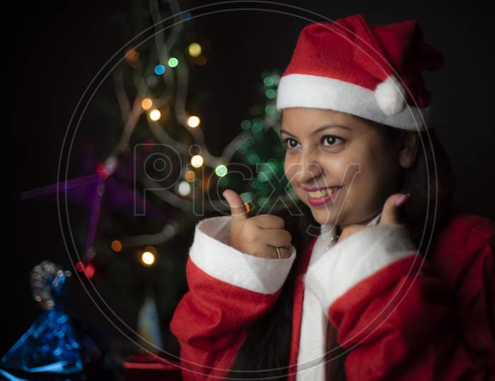 Close up portrait of young and cheerful Indian Bengali plus size brunette woman in Santa Clause jacket and hat in decorative Christmas tree background.Indian lifestyle and Christmas celebration.