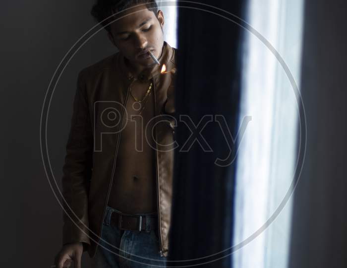 Close up portrait of an young and handsome man with front open leather jacket lightening cigarette while standing in front of a glass door of a balcony. Indian lifestyle.