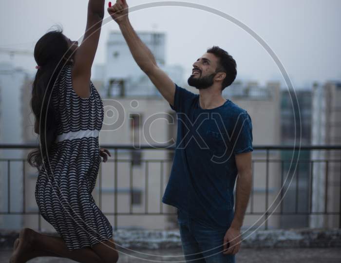 A dark skinned Indian/African girl in  western dress jumping to take the umbrella from her kashmiri/European/Arabian boyfriend on a rooftop in afternoon in urban background. Lifestyle of a couple.