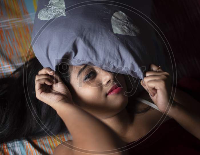 An young and attractive Indian Bengali brunette woman in white sleeping wear is lying on a bed with a blanket in her room in colorful background. Indian lifestyle.