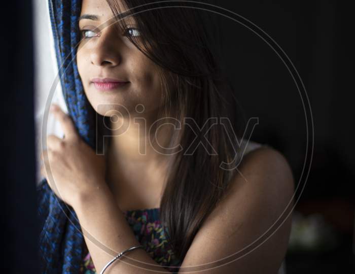 An young and attractive Indian brunette woman in white sleeping wear looking outside through the window removing the curtain in dark background. Indian lifestyle.