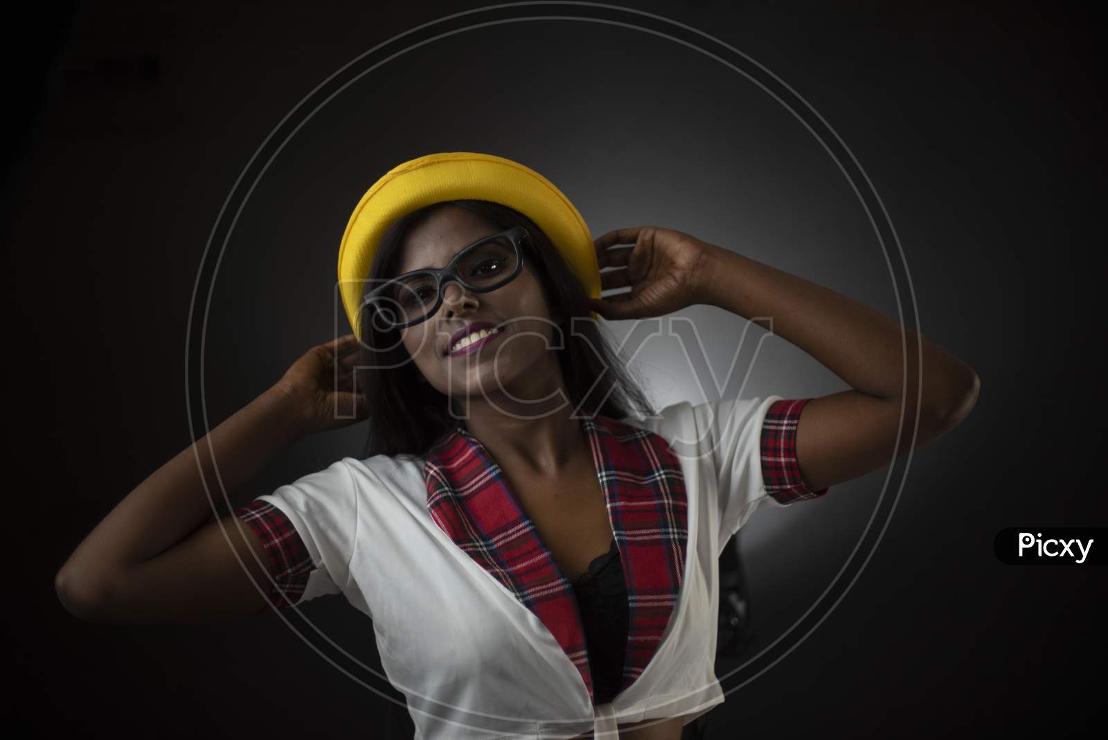 Image of Portrait of an Asian/Indian/African brunette dark skinned young  girl in sexy school uniform and spectacles with yellow hat in a black/grey  studio background. Fashion and cosplay photography.-QG883078-Picxy