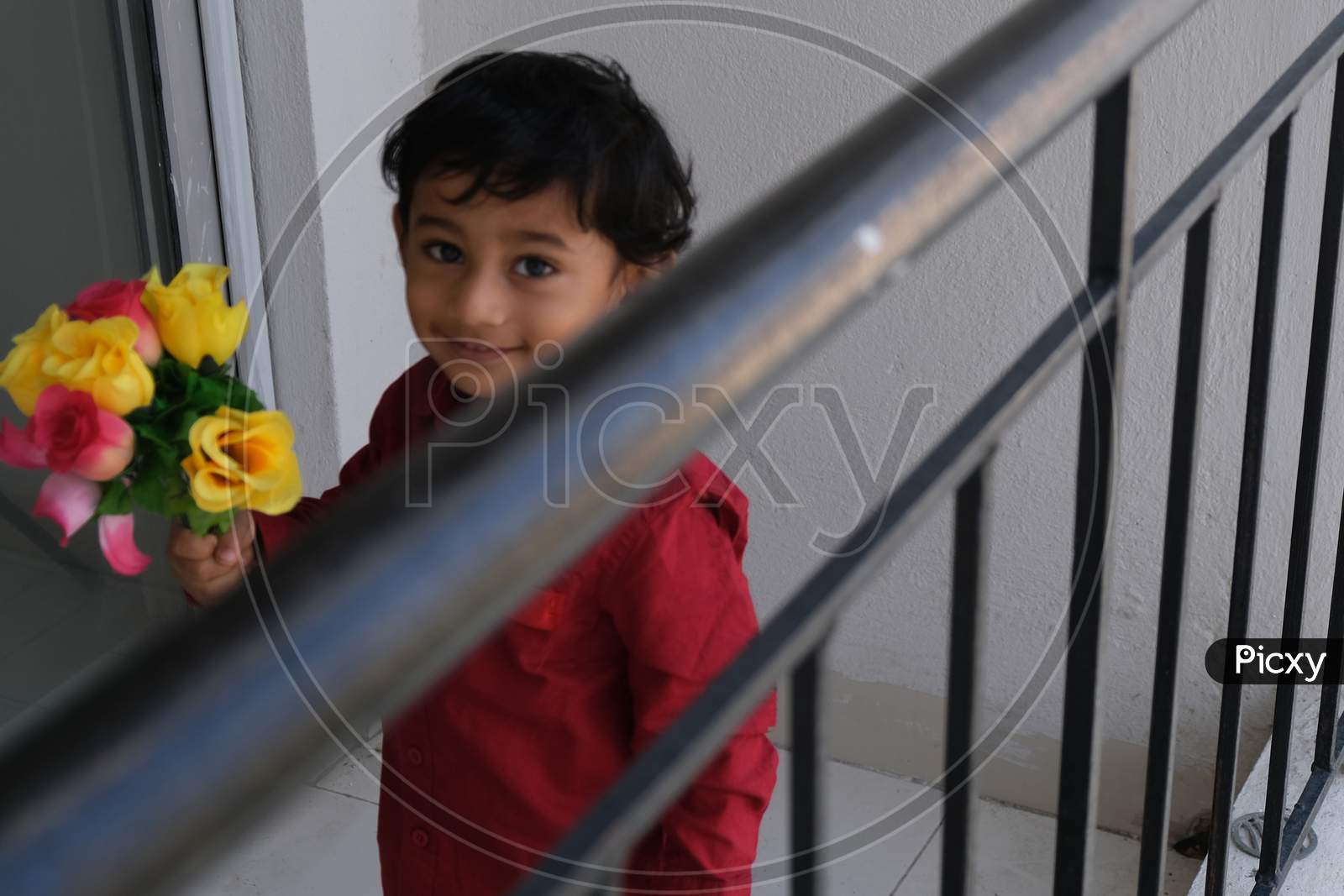 Portrait of Indian cute little cheerful brunette Tamil baby boy wearing vibrant red shirt while standing on a balcony in a white background. Indian lifestyle.