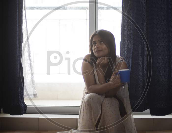 An young and attractive Indian  brunette woman in white sleeping wear in front of a balcony window with a tea/coffee mug in the morning in white background. Indian lifestyle.