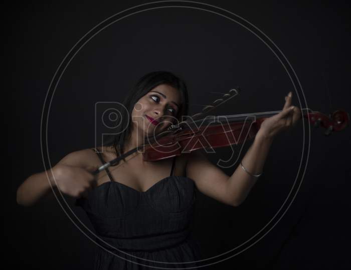 Fashion portrait of an young and attractive Indian Bengali brunette girl with black western dress is playing violin in front of a black studio background. Indian fashion portrait and lifestyle.