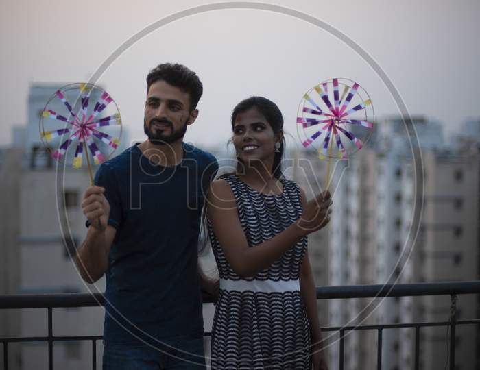 A dark skinned Indian/African girl in  western dress and a Kashmiri/European/Arabian man in casual wear playing with pinwheel on a rooftop in urban background. Indian lifestyle.