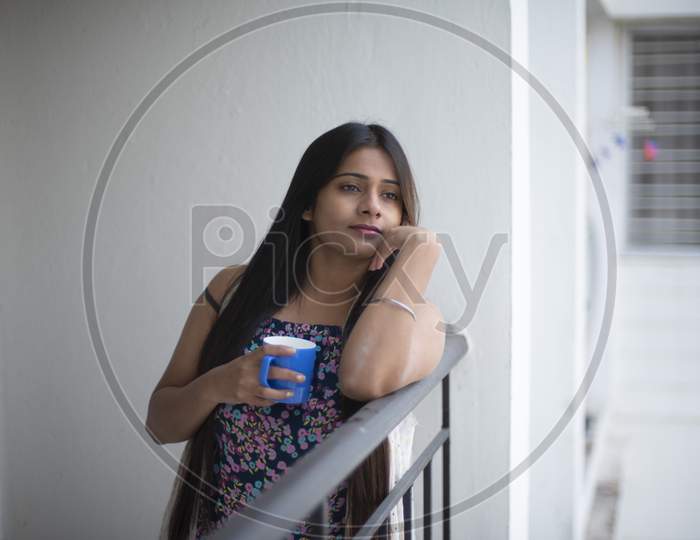 An young and attractive Indian brunette woman in white sleeping wear standing in a balcony with a tea/coffee mug in the morning in white background. Indian lifestyle