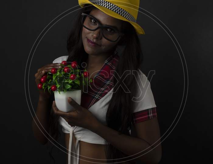 Portrait of Asian/Indian/African brunette dark skinned young girl in sexy school uniform and spectacles with yellow hat holding flower in black/grey studio background. Fashion and cosplay photography.