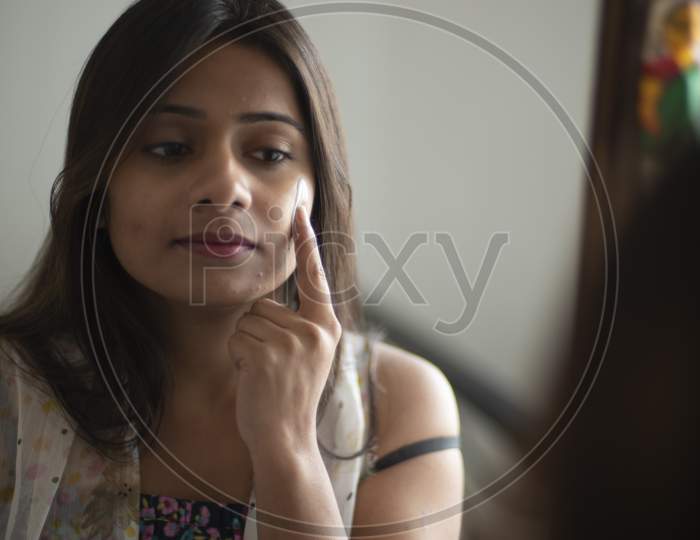 Reflection of an young and attractive Indian Bengali brunette woman is looking her face in a mirror while applying a white beauty cream on her face in her room. Indian lifestyle.