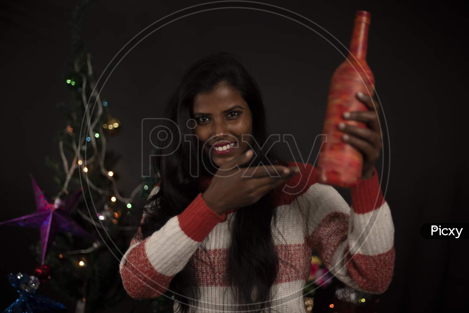 Portrait of an young and attractive brunette dark skinned Indian Bengali woman in western woolen sweater showing gift in decorative Christmas tree background.Indian lifestyle and Christmas celebration