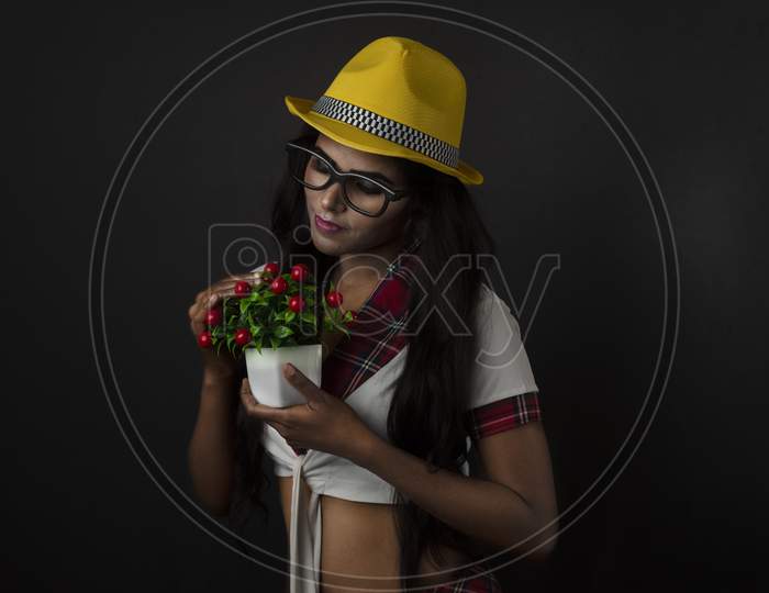 Portrait of Asian/Indian/African brunette dark skinned young girl in sexy school uniform and spectacles with yellow hat holding flower in black/grey studio background. Fashion and cosplay photography.