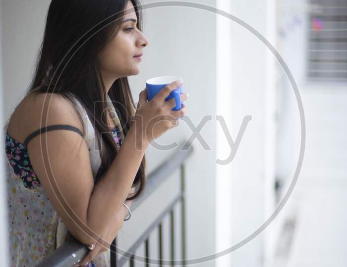 An young and attractive Indian brunette woman in white sleeping wear standing in a balcony with a tea/coffee mug in the morning in white background. Indian lifestyle