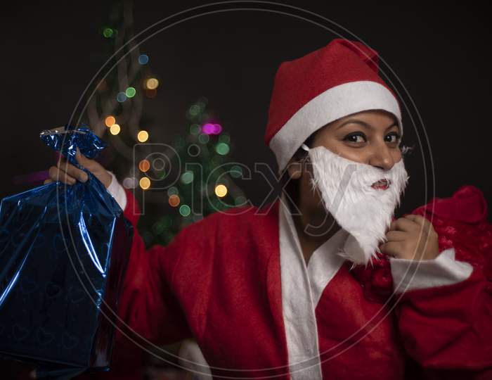 Young Indian plus size brunette woman in Santa Clause jacket and hat carrying her red bag in a funny and happy mood in decorative Christmas tree background. Indian lifestyle and Christmas celebration.