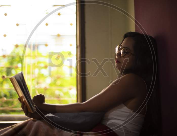 A young and attractive Indian Bengali brunette woman in white sleeping wear with a coffee/tea cup reading book on a bed in front of a window inside in her room. Indian lifestyle.
