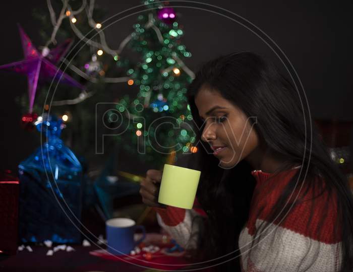 An young and attractive brunette dark skinned Indian Bengali woman in western woolen sweater enjoying with coffee/tea in decorative Christmas tree background.Indian lifestyle and Christmas celebration