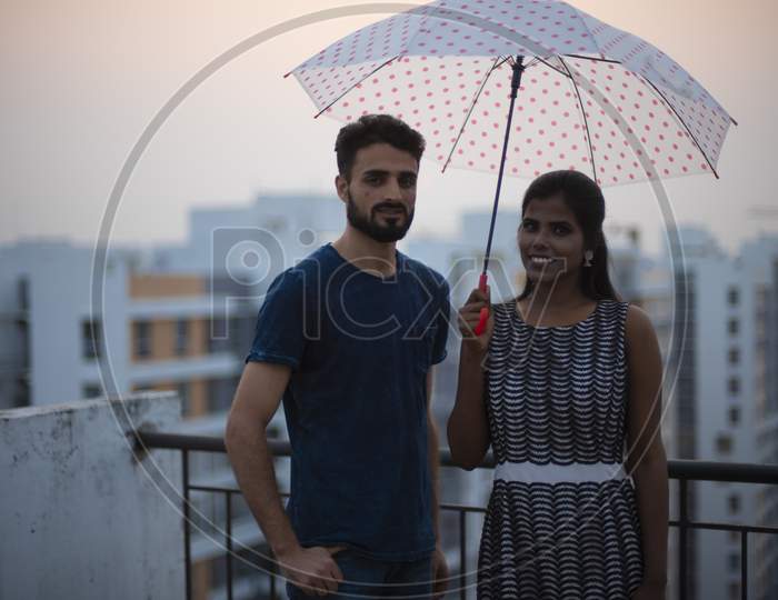 A dark skinned Indian/African girl in  western dress and a Kashmiri/European/Arabian man in casual wear having fun with umbrella on a rooftop in urban background. Indian lifestyle.