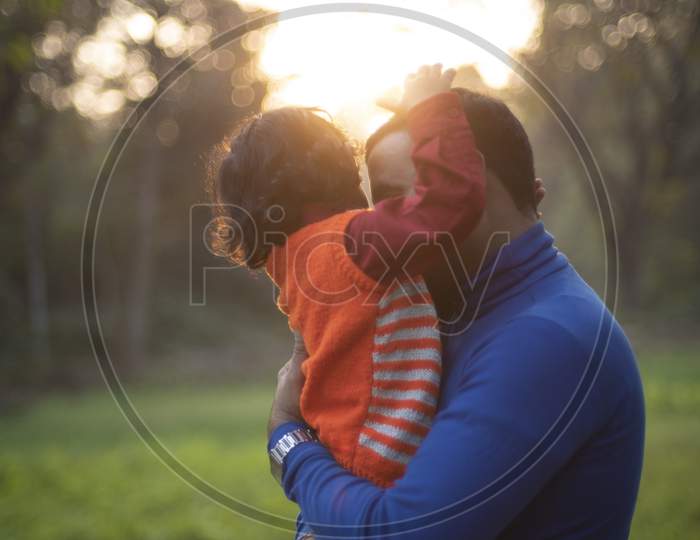 An Indian Telugu brunette father and his baby boy in winter garments enjoying themselves in winter afternoon on a  green grass field in forest background. Indian lifestyle and back light photography.