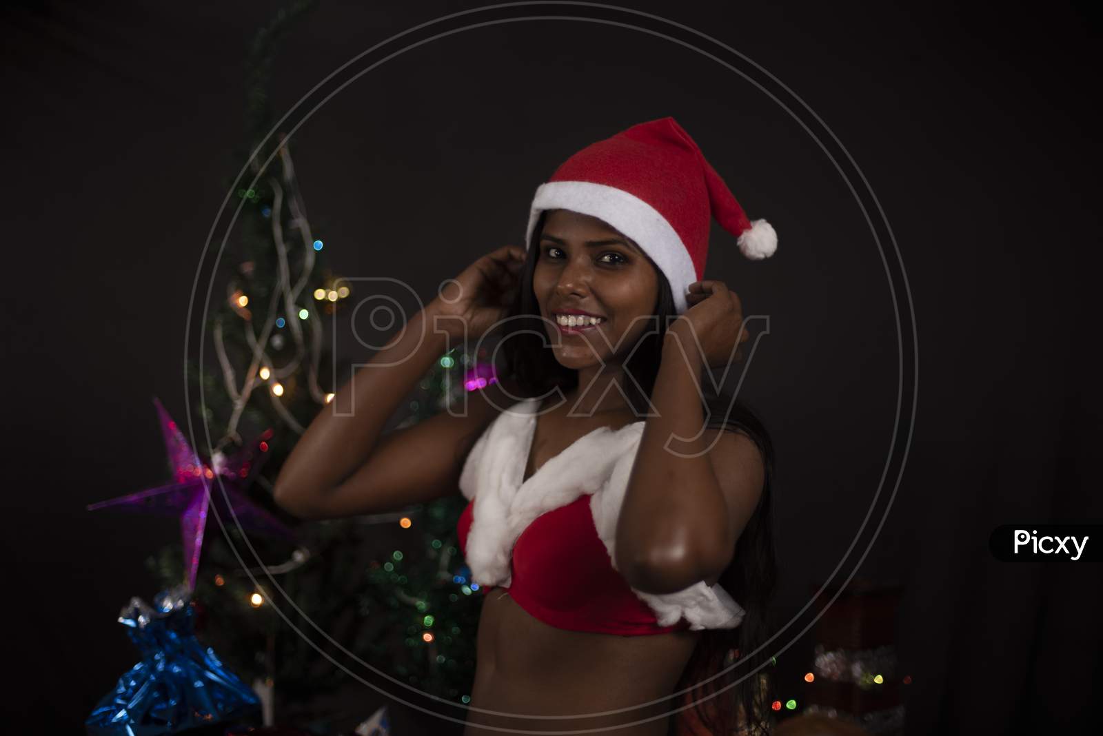 Portrait of young Indian Bengali dark skinned brunette woman in Santa Clause lingerie, jacket and hat with gifts in decorative Christmas tree background.Indian lifestyle and Christmas celebration.