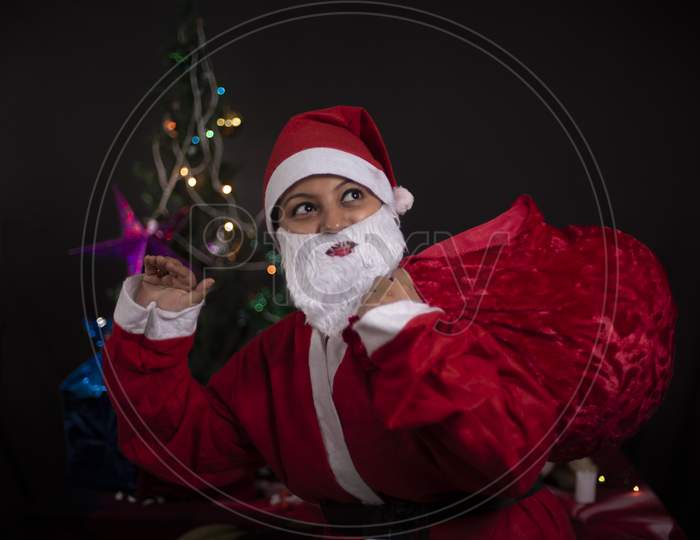 Young Indian plus size brunette woman in Santa Clause jacket and hat carrying her red bag in a funny and happy mood in decorative Christmas tree background. Indian lifestyle and Christmas celebration.