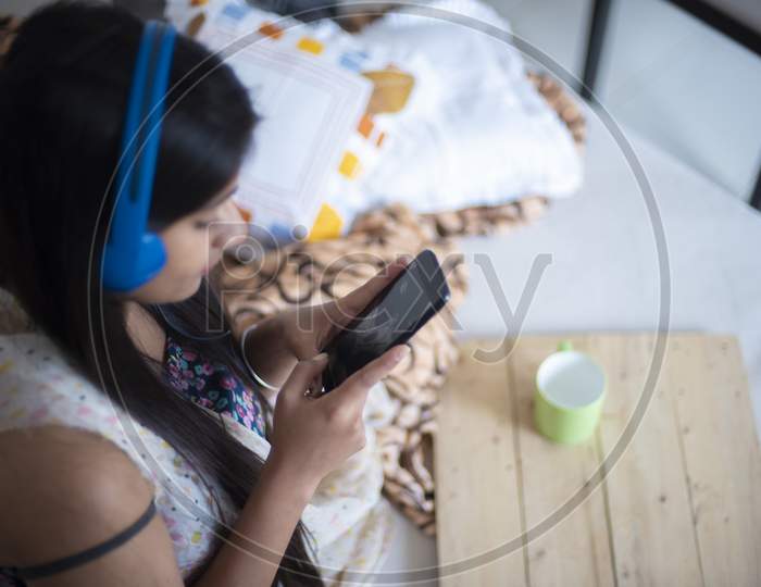 An young Indian Bengali brunette girl in white sleeping wear is listening music while sitting on bed with cellphone,coffee/tea cup and earplug in a casual mood in the winter morning. Indian lifestyle.
