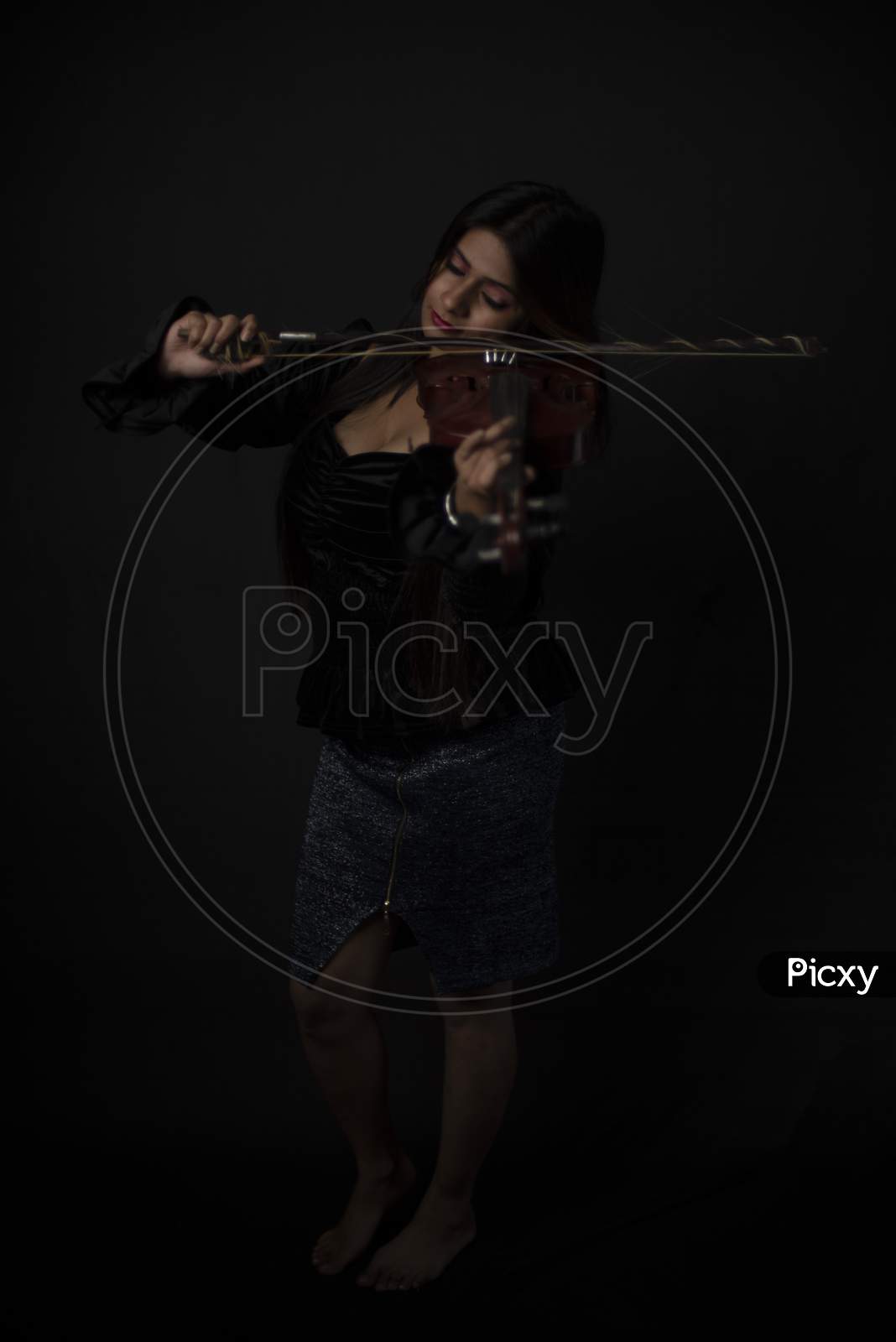 Fashion portrait of an young and attractive Indian Bengali brunette girl with black velvet dress is playing violin in front of a black studio background. Indian fashion portrait and lifestyle.