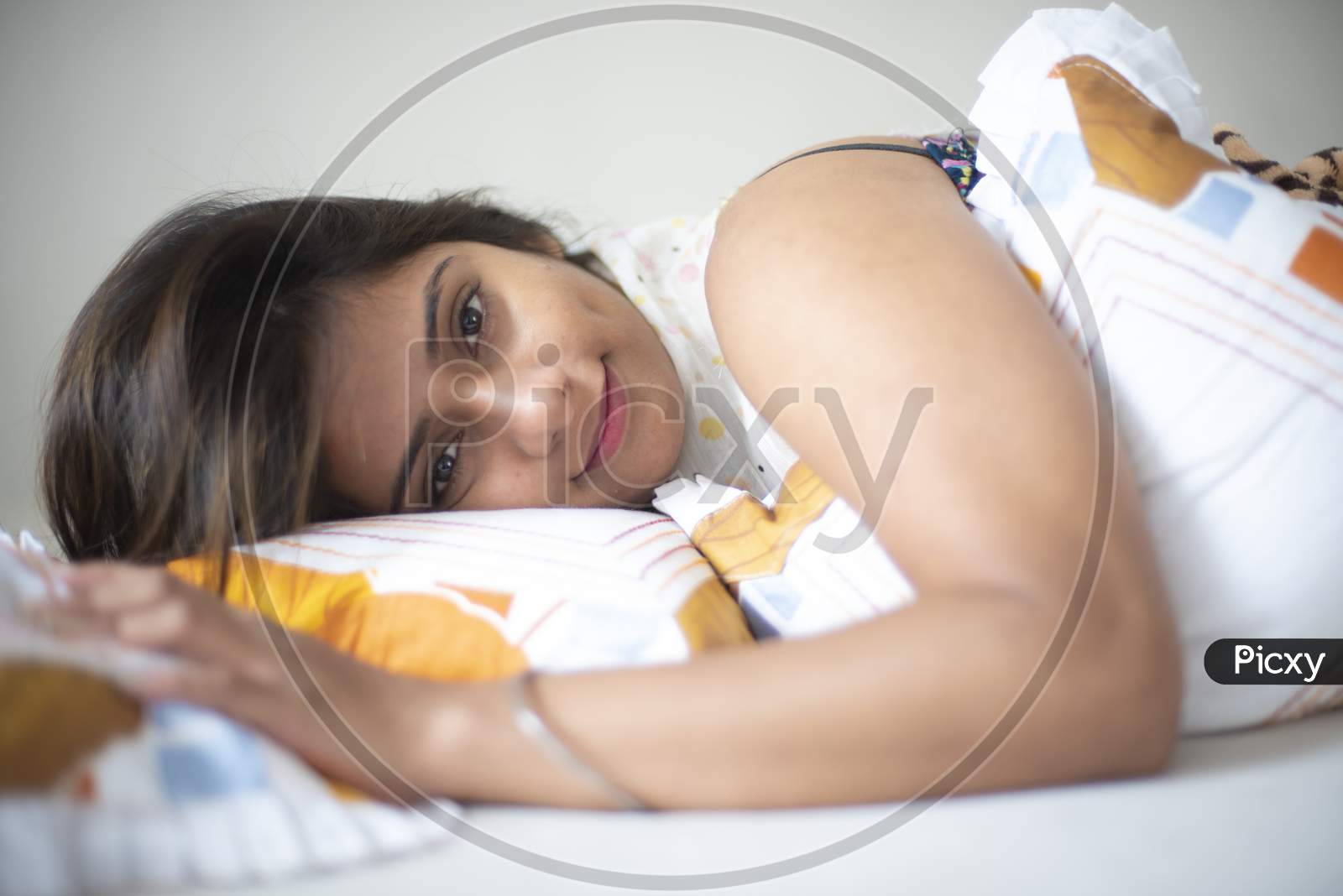 Top down portrait of an young and attractive Indian Bengali brunette woman in white sleeping wear is lying on a white bed with white cushions and blanket in her bedroom. Indian lifestyle.
