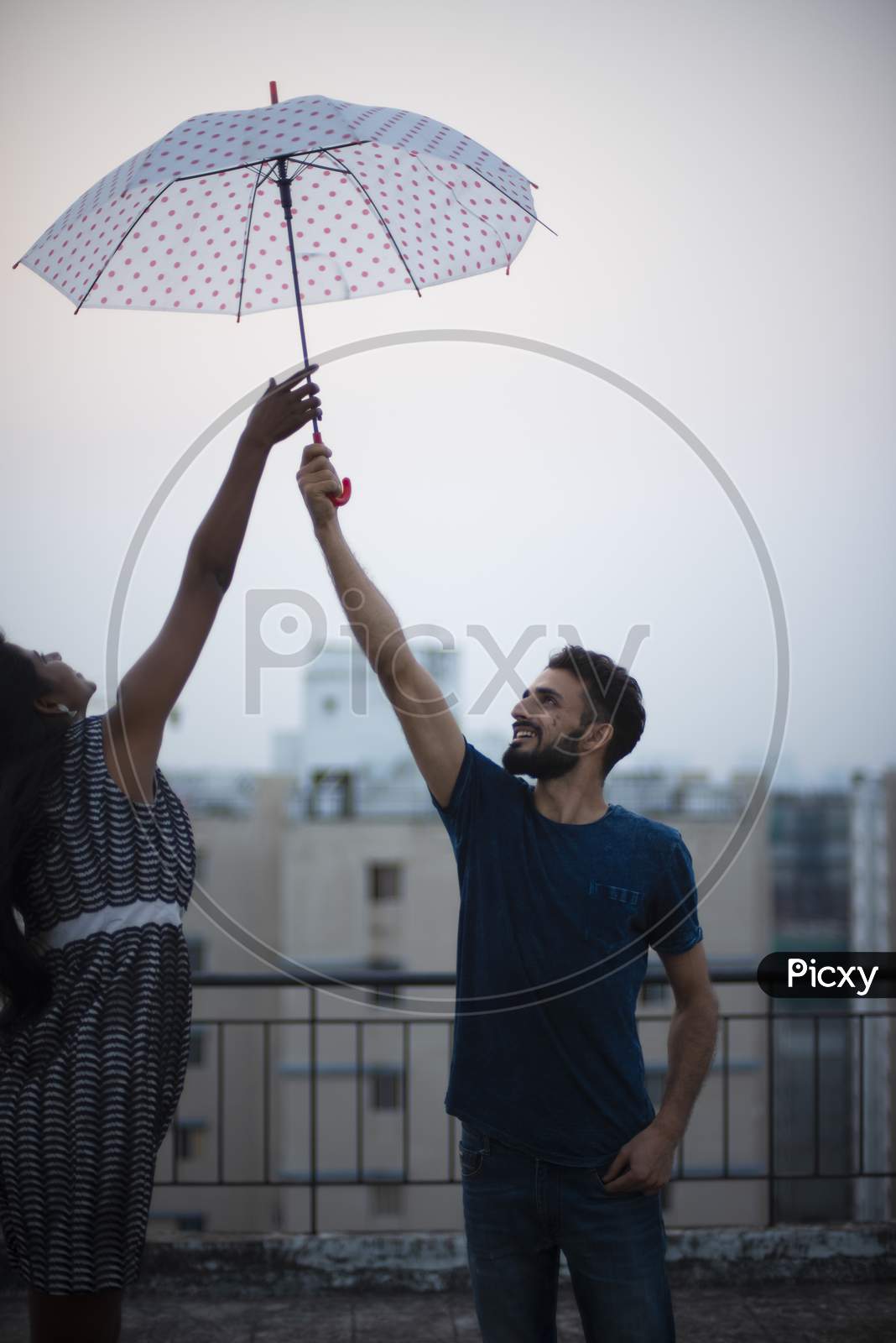 A dark skinned Indian/African girl in  western dress jumping to take the umbrella from her kashmiri/European/Arabian boyfriend on a rooftop in afternoon in urban background. Lifestyle of a couple.