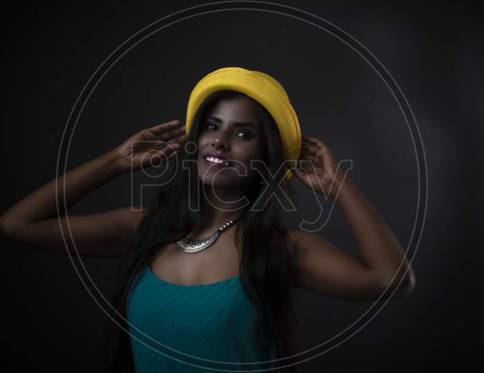 Fashion portrait of young dark skinned Indian/African brunette girl in green western dress, jewelry and yellow hat in front of black copy space studio background. Indian fashion photography.