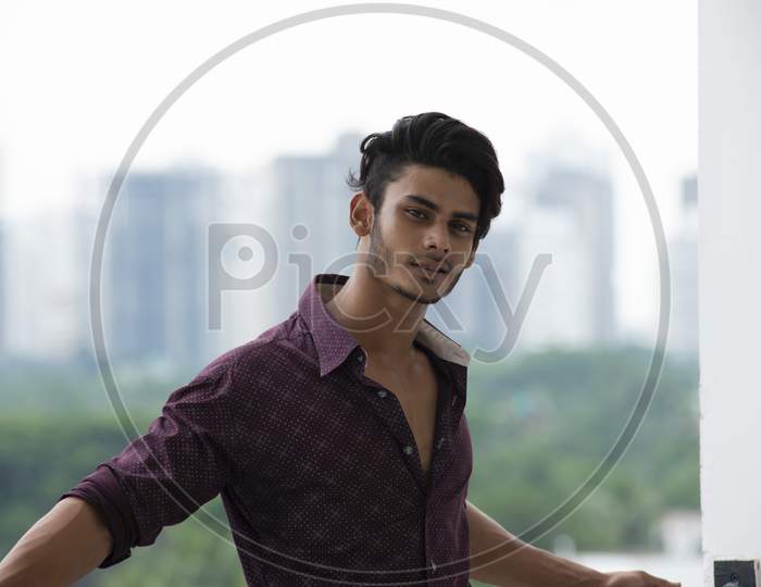 Portrait of an young and handsome brunette Bengali muscular man in casual shirt and jeans standing on a balcony in white urban background. Indian lifestyle.
