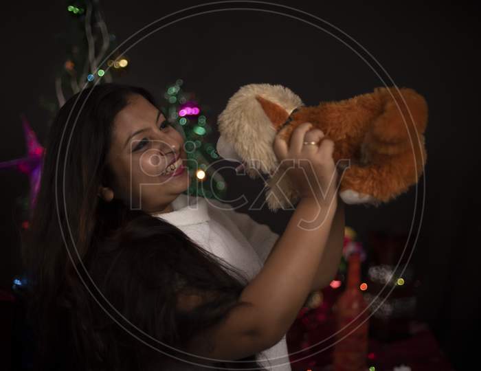 An young and attractive brunette plus size Indian Bengali woman in western woolen sweater enjoying with soft toy  in decorative Christmas tree background.Indian lifestyle and Christmas celebration