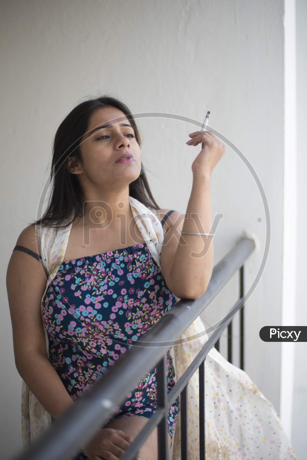 An young and attractive Indian Bengali brunette woman in white sleeping wear is smoking cigarette on the balcony in the morning in white background. Indian lifestyle.